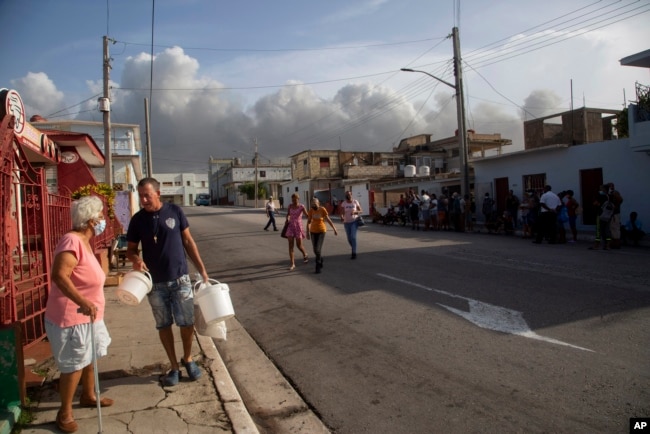 Residentes caminan por una calle de Matanzas este 9 de agosto, mientras se eleva al fondo el humo del incendio en la base de almacenamiento de crudo. (AP Foto/Ismael Francisco)