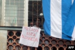 Oficialistas colgaron banderas cubanas en las ventanas de la casa de Yunior García, en un intento de evitar que se comunique con el exterior, mientras sostiene una rosa blanca.